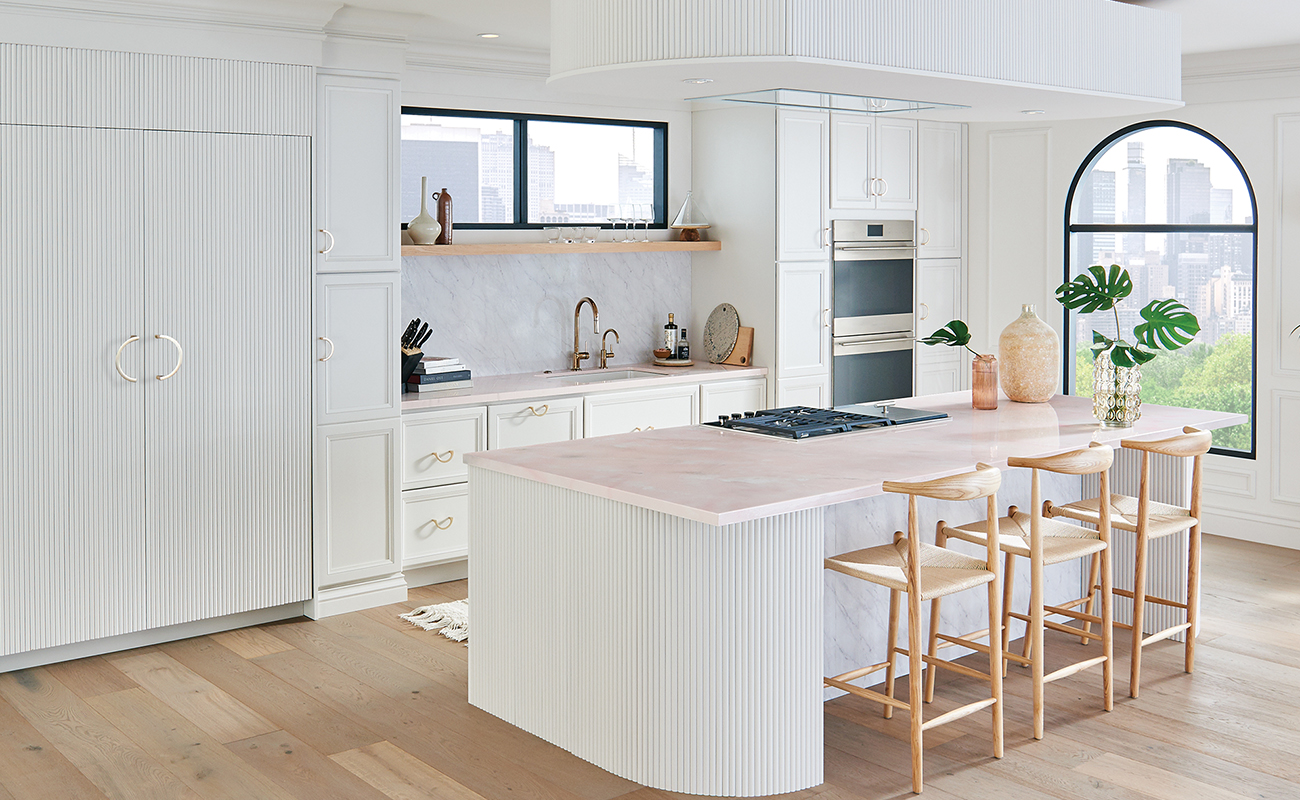 A bright, all-white kitchen with an elegant flat panel cabinet door style and unique reeded details with a curved kitchen island and dropdown ceiling feature.