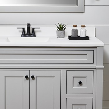 A grey and white bathroom with a light grey painted vanity with inset cabinet construction and beautiful furniture styled details. A white painted shiplap wall, a matching light gray framed mirror, and a solid white vanity top pull this look together.