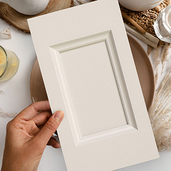 A flat lay of a person holding a cabinet drawer front sample with a deep center panel and a soft, natural white paint finish.