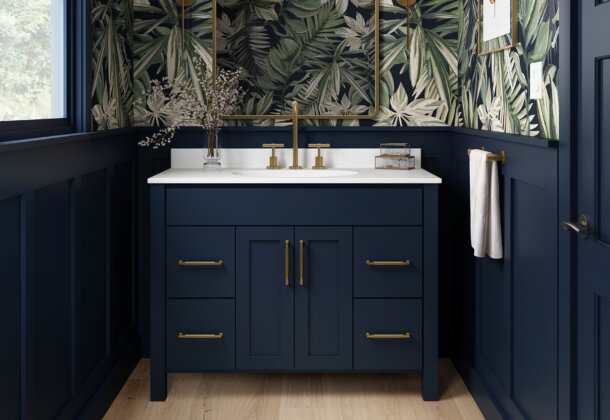 A trendy navy blue bathroom with a furniture style blue vanity with a solid white countertop, gold/brushed brass hardware, blue wainscot panels, and a fashionable blue, green, and white wallpaper.