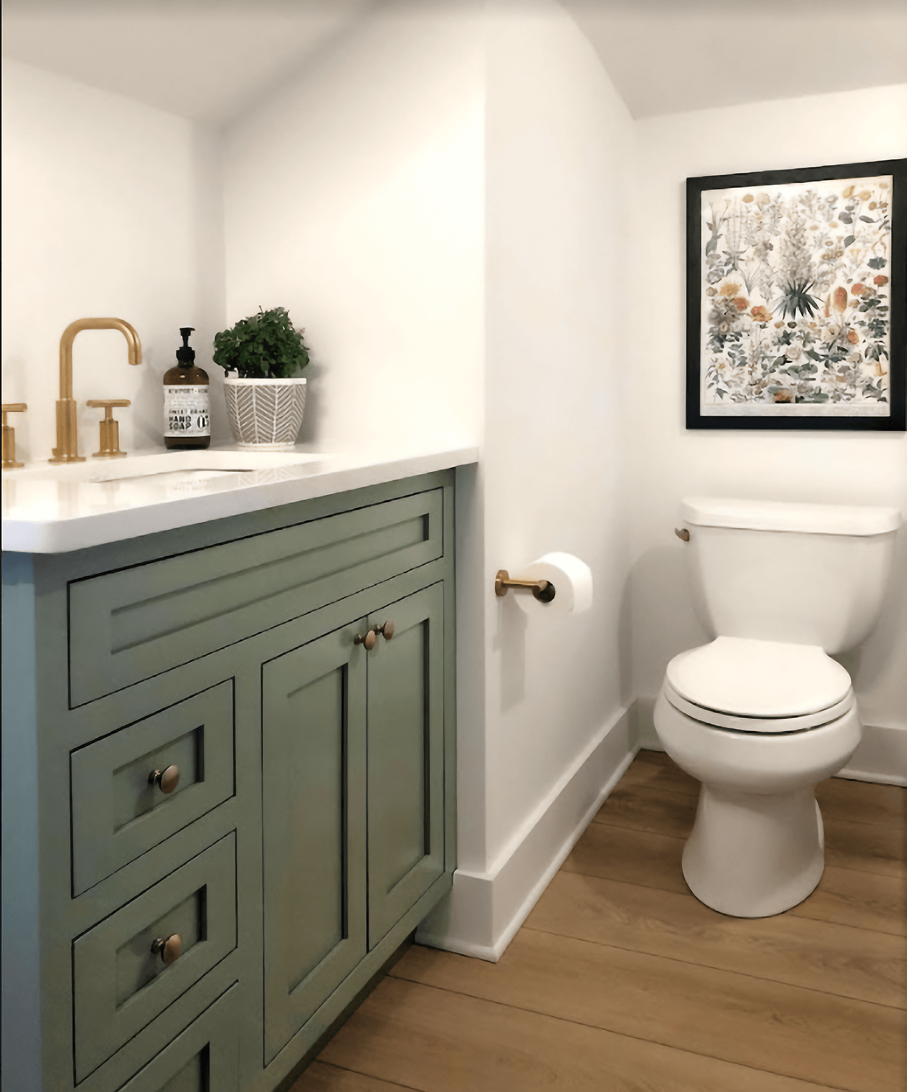 A soft, sage green painted bathroom vanity with black cabinet hardware, a solid white vanity top, and a brushed brass faucet.